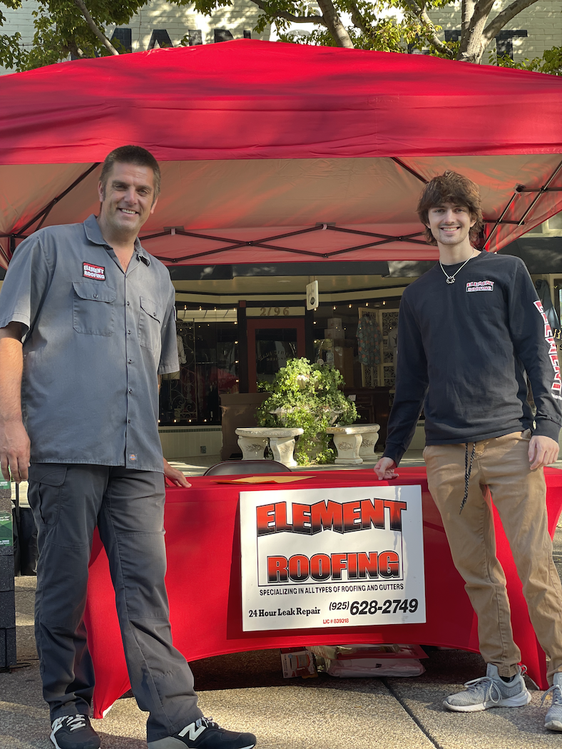 Chad Maimone and his son standing at an Element Roofing information booth under a red canopy tent at a street fair in Pleasanton, California.
